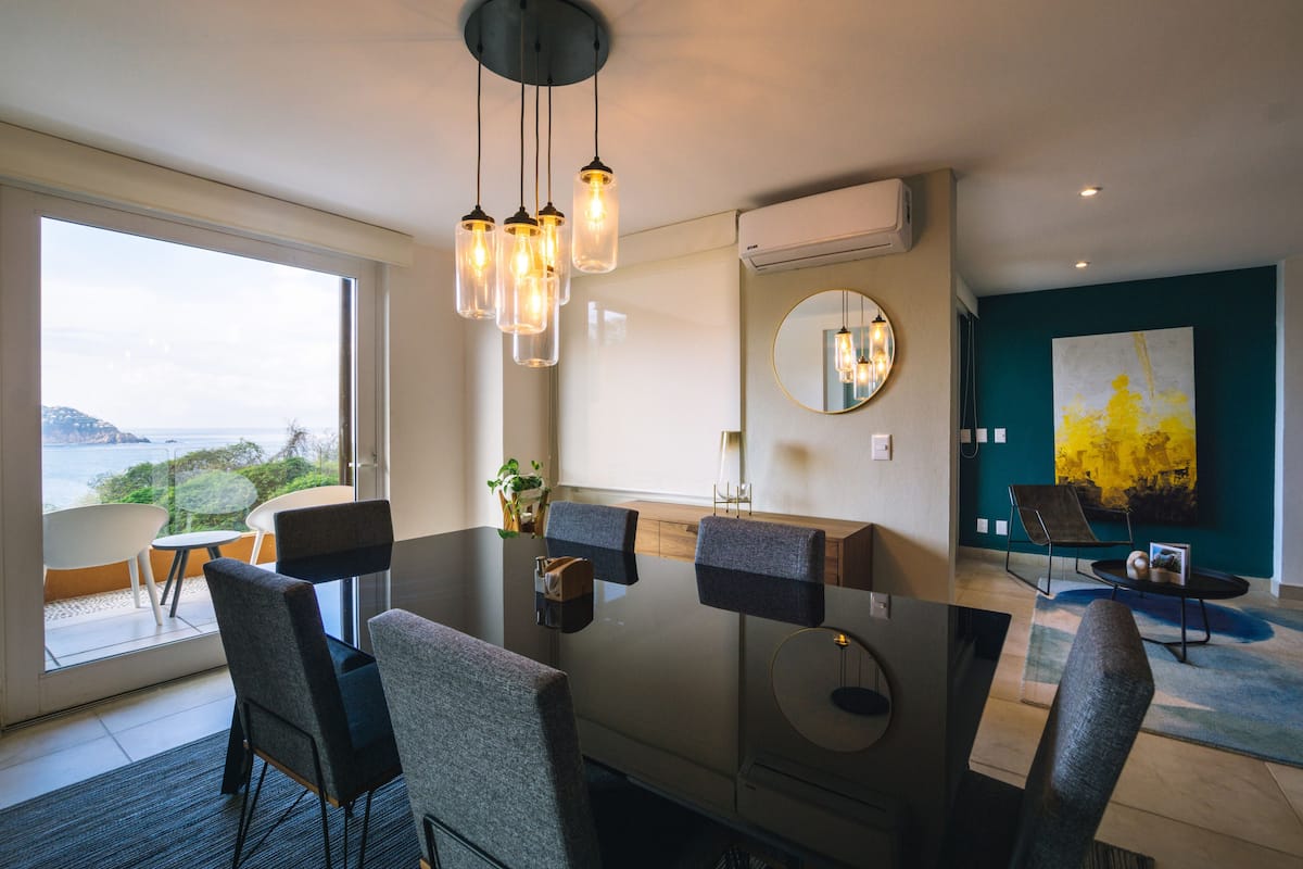 Un comedor moderno con una mesa de vidrio, varias sillas, iluminación colgante, vistas al exterior y un cuadro grande en la pared.