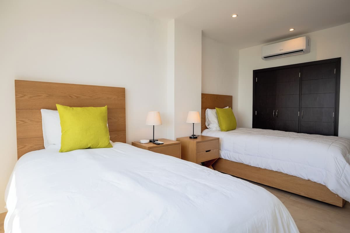 Habitación de hotel con dos camas, cada una con cabecero de madera y ropa de cama blanca. Se observan mesas de noche con lámparas y un armario grande.