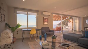 Ein modernes Wohnzimmer mit einer Couch, einem Glastisch, einem Sessel und Blick ins Freie durch große Fenster.