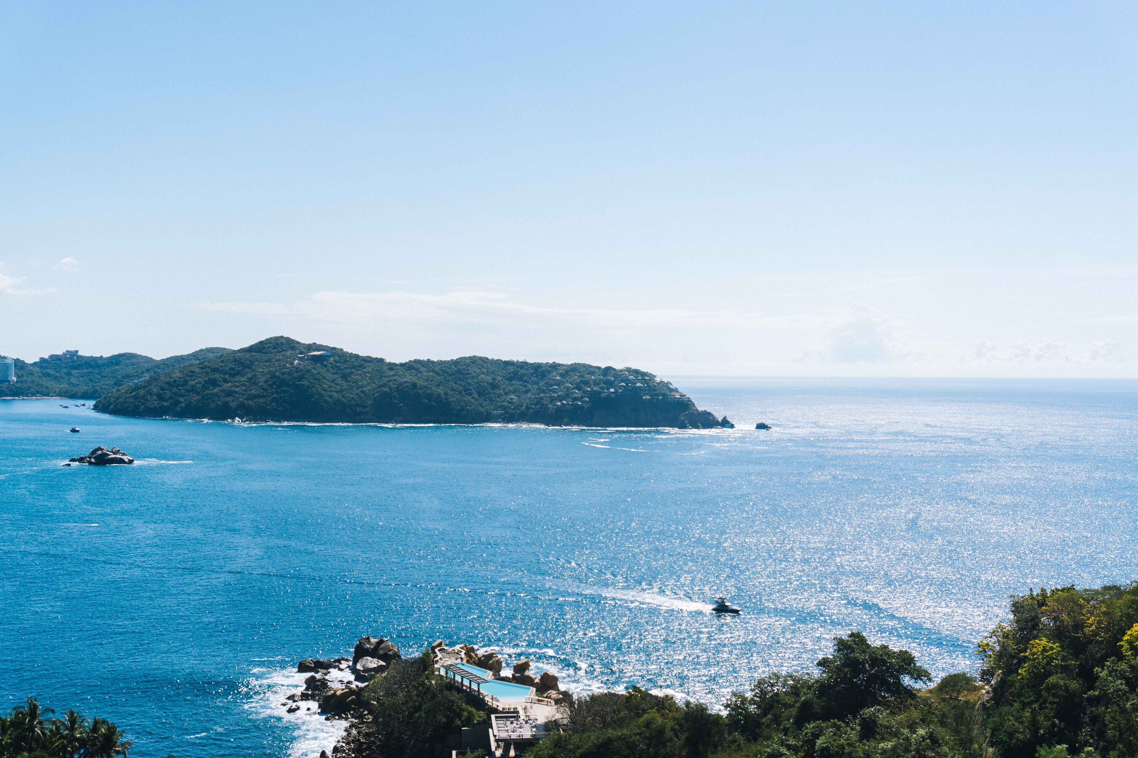 Vista costeira com céu claro, uma pequena ilha e um barco na água.
