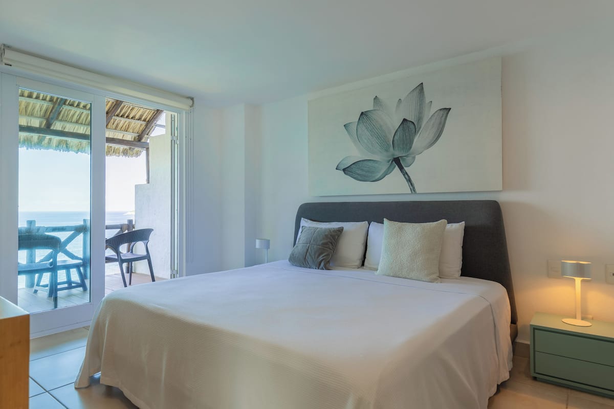 Un dormitorio con cama, mesita de noche, lámpara, un cuadro de una flor y vistas al exterior.