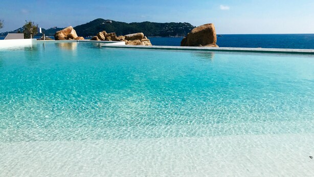 Ein klares, blaues Schwimmbecken mit großen Steinen und Blick auf das Meer sowie Berge im Hintergrund.