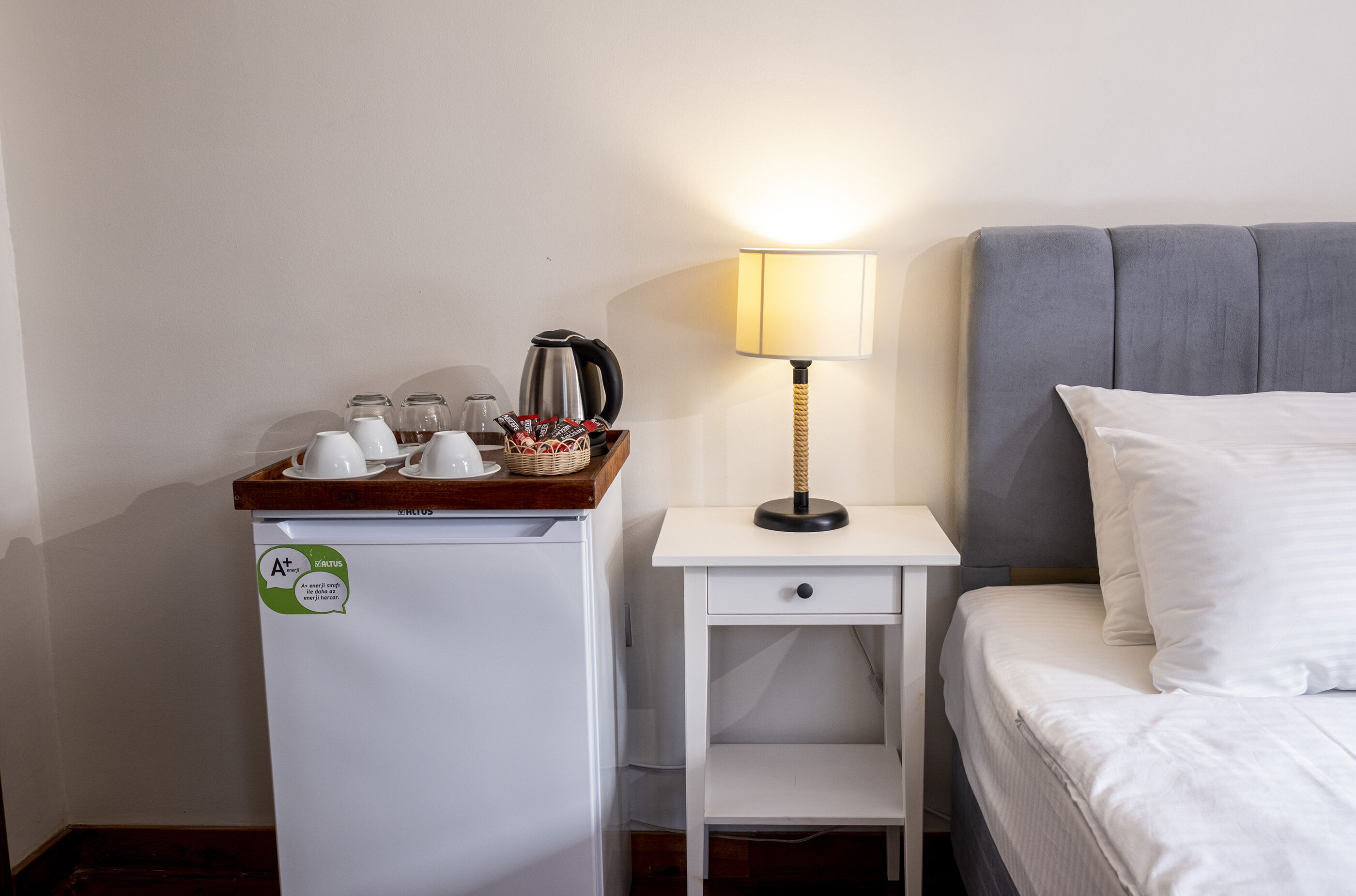 A compact hotel room with a bed, a small bedside table with a lamp, a mini fridge with a coffee maker, and a basket of snacks.