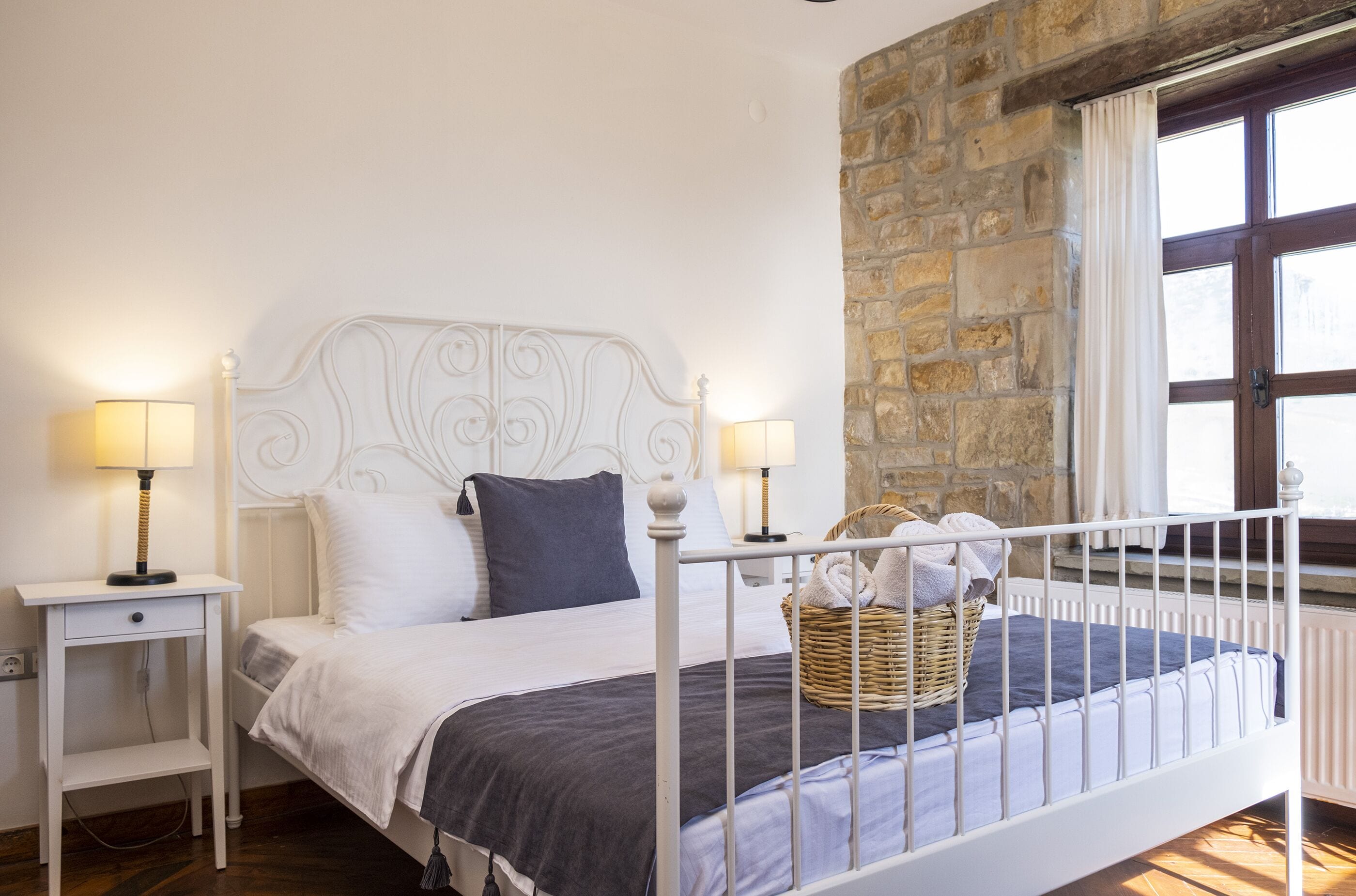 A bed with a white metal frame, two bedside tables with lamps, a stone wall, and a window with curtains.