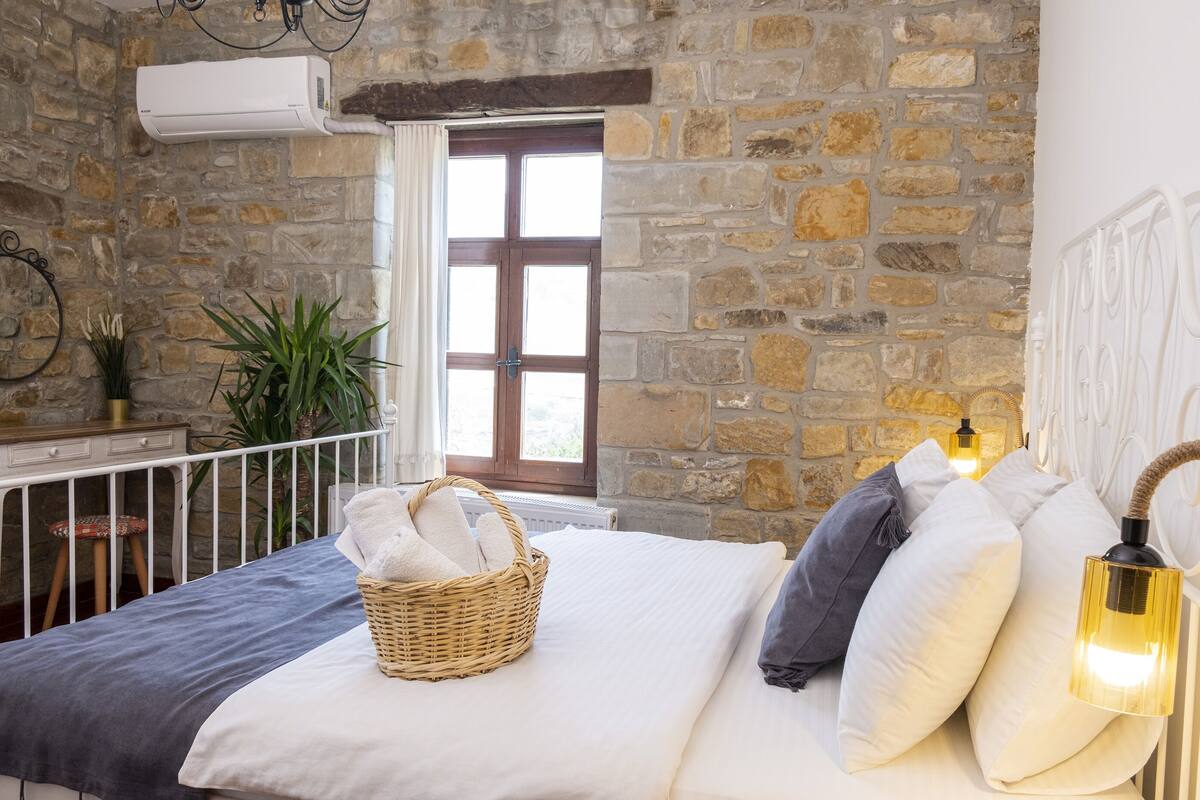 Quarto com parede de pedra, janela com persianas de madeira, cama com roupa de cama branca, cesto de vime com toalhas e abajur.