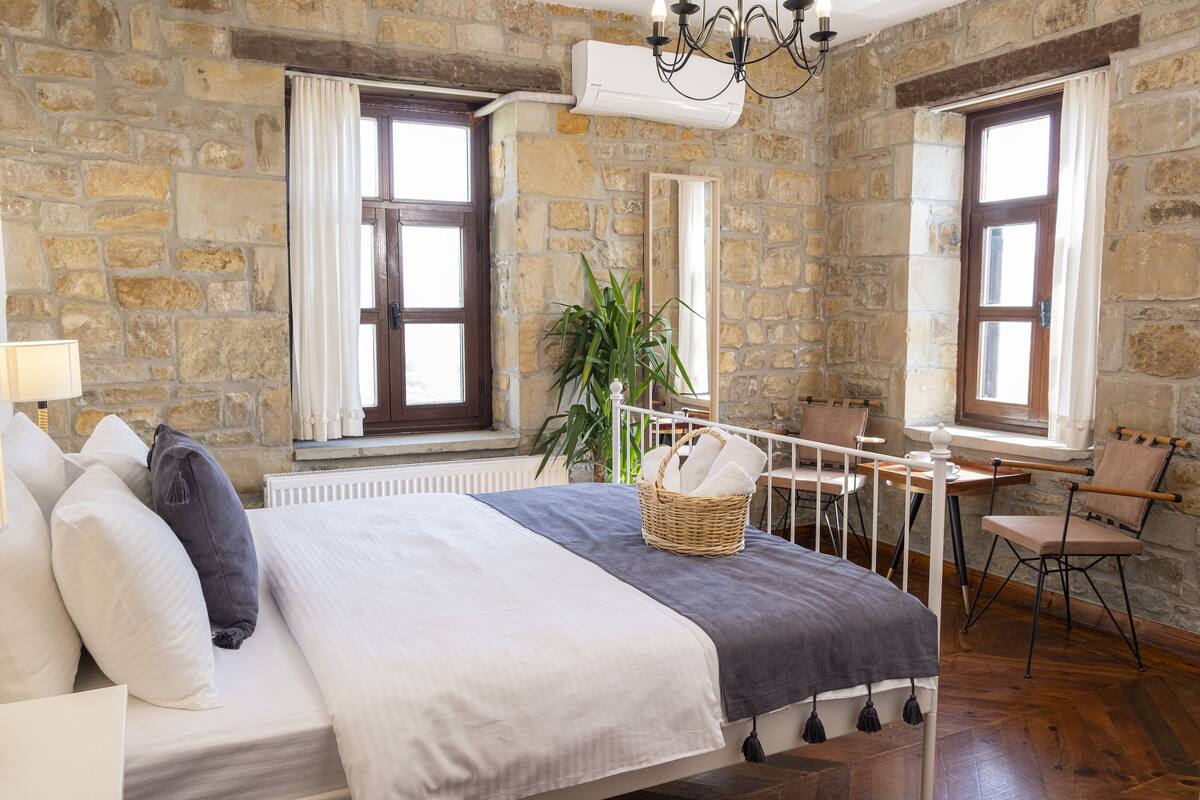 Quarto com parede de pedra, cama com roupa de cama branca e cinza, uma cadeira, uma pequena mesa e uma planta.