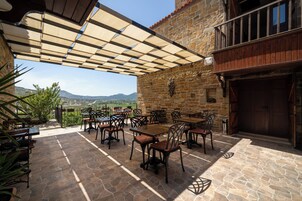 Un patio al aire libre con suelo de piedra, mesas y sillas, un balcón de madera y un muro de piedra con una decoración de vid.
