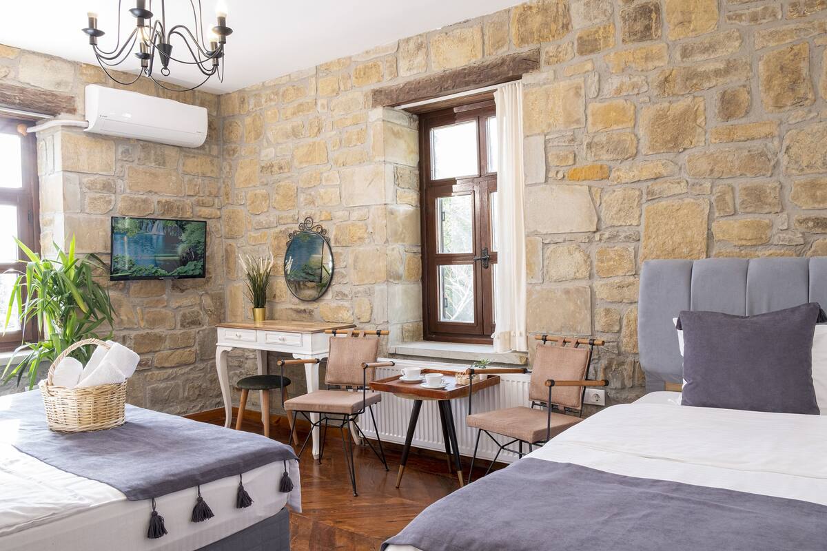 Um quarto aconchegante com parede de pedra, uma cama, escrivaninha com cadeiras e uma televisão.