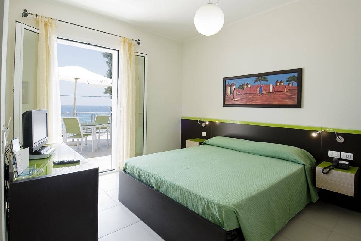 A hotel room with a bed, a desk with a computer, a chair, a phone, and a painting on the wall.