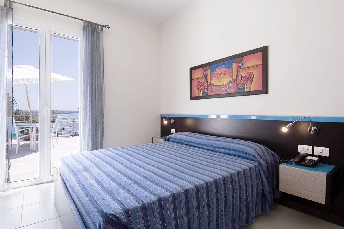 A hotel room with a bed, a nightstand with a phone, a painting on the wall, and a view through a sliding door to an outdoor area with a table and chairs.