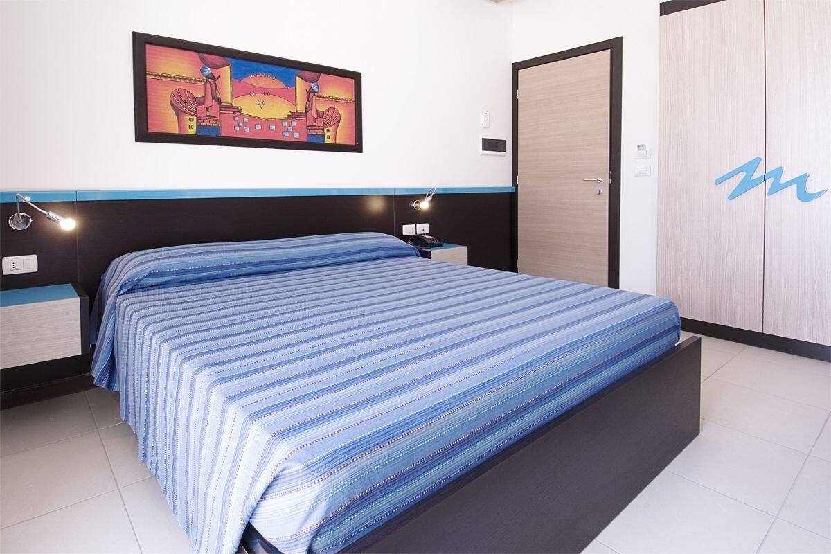 A bedroom with a bed, a nightstand, a wall-mounted lamp, a door, and a painting.