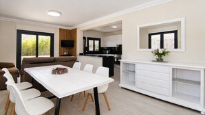 A modern living room with a white dining table, a sofa, and a kitchen area with white cabinetry and a flat-screen TV. - Ca Na Coloma 145 fantastic villa with barbecue, billiards, ping pong and WiFi (Playa de Muro)