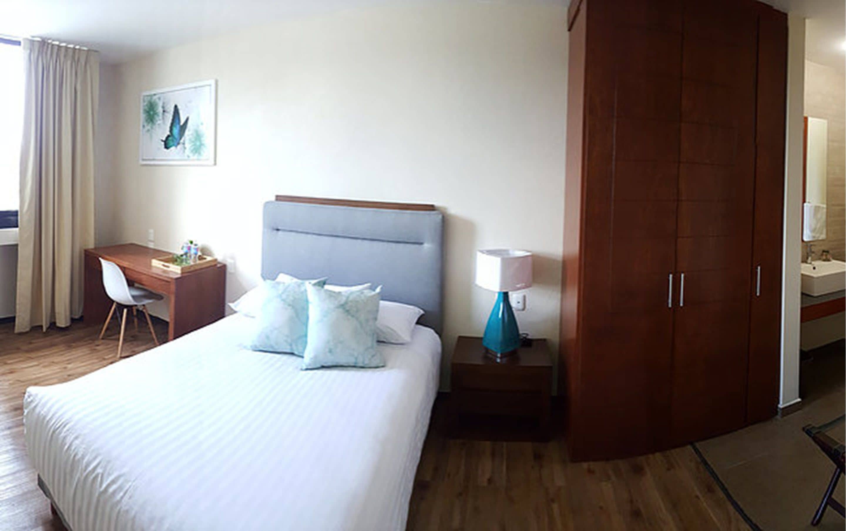 Una habitación de hotel con una cama, un escritorio, una silla, una mesita de noche con lámpara, un armario y un cuadro en la pared.
