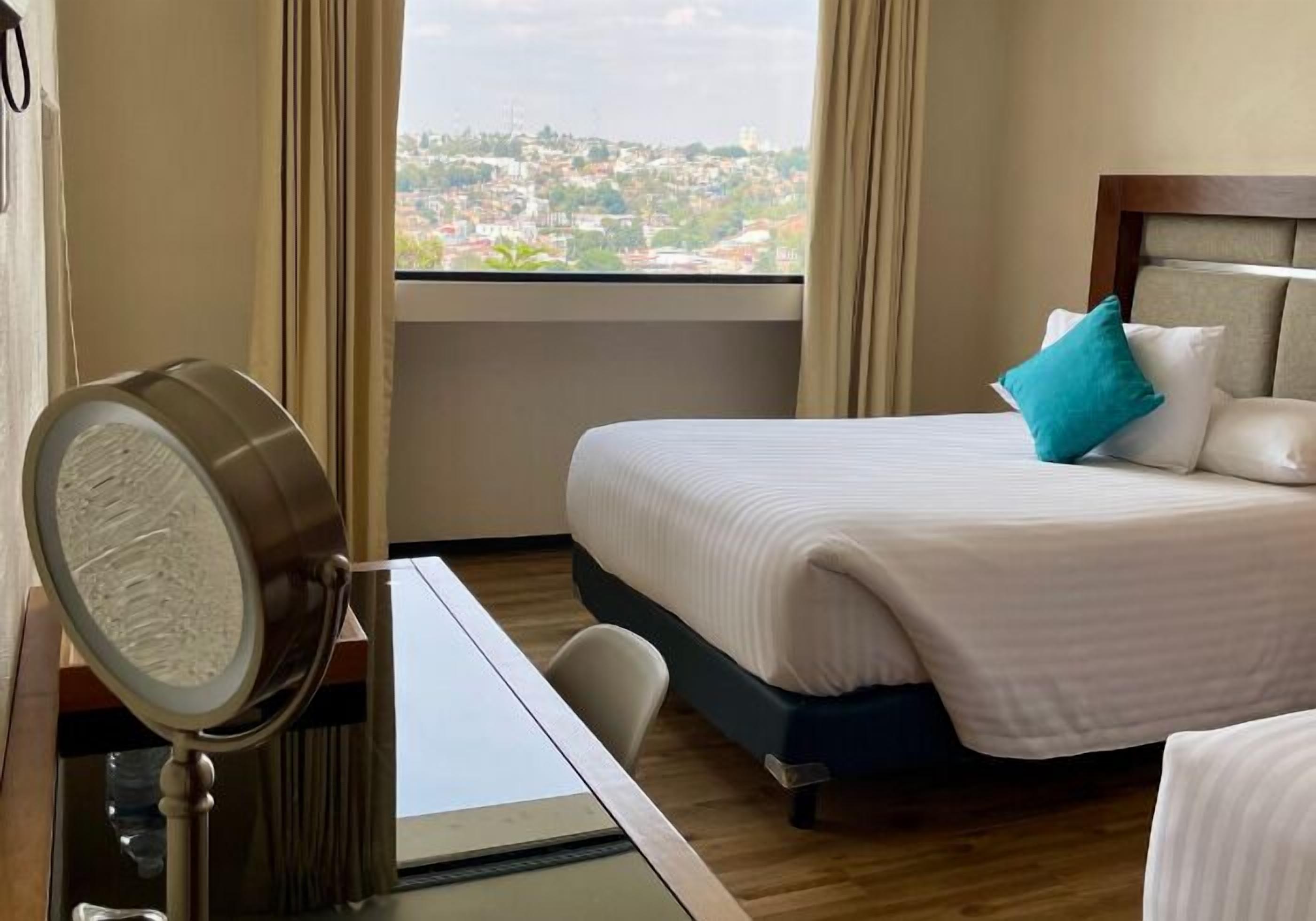 A hotel room with a bed, a desk, a chair, and a view of a cityscape through the window.