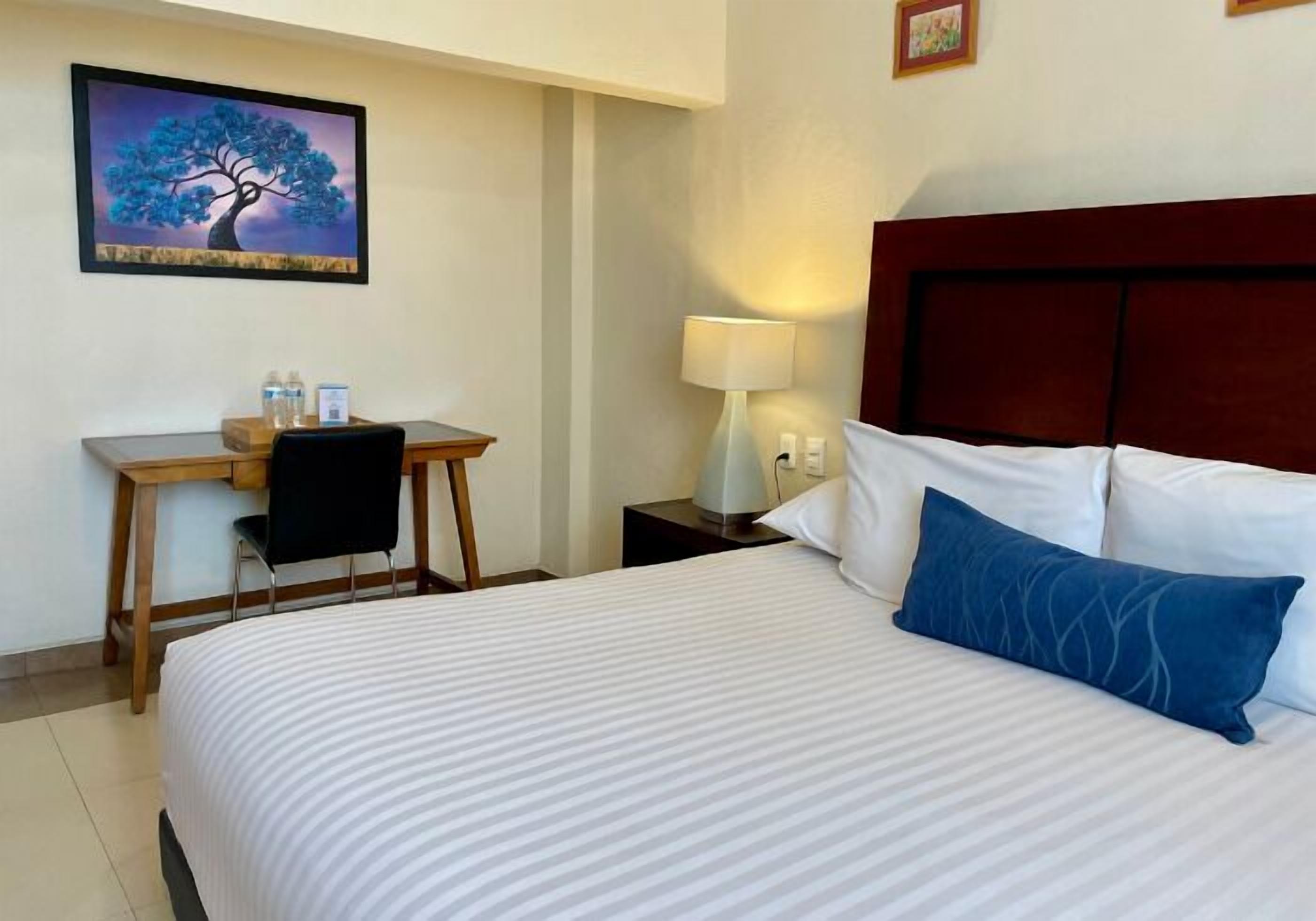 Habitación de hotel con cama, escritorio, silla, lámpara y un cuadro en la pared.