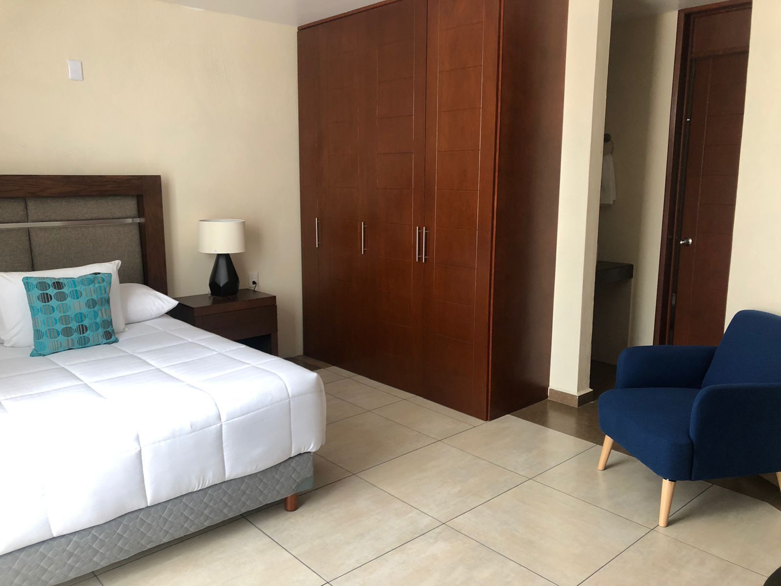 Una habitación de hotel con cama, mesita de noche con lámpara, armario y sillón azul.