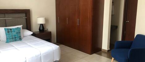 Una habitación de hotel con cama, mesita de noche con lámpara, armario y sillón azul.