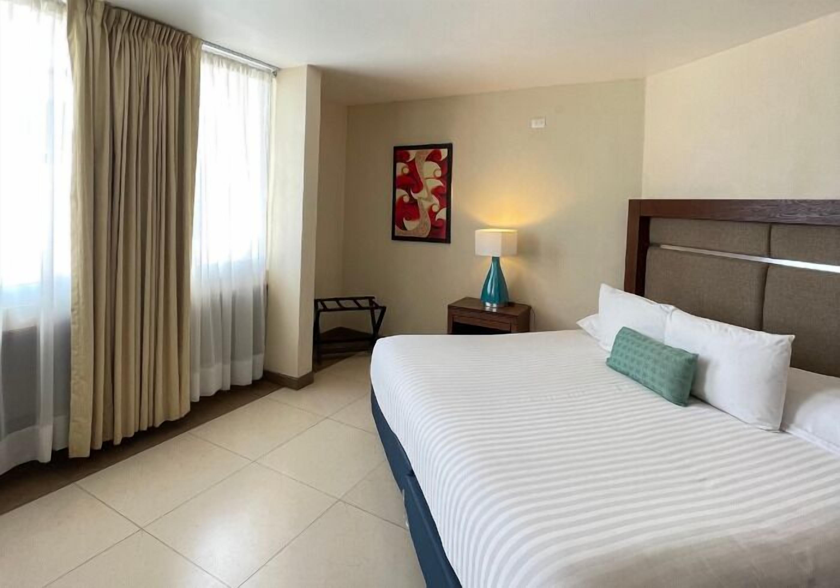 A hotel room with a large bed, a nightstand with a lamp, a framed picture on the wall, and a window with curtains.