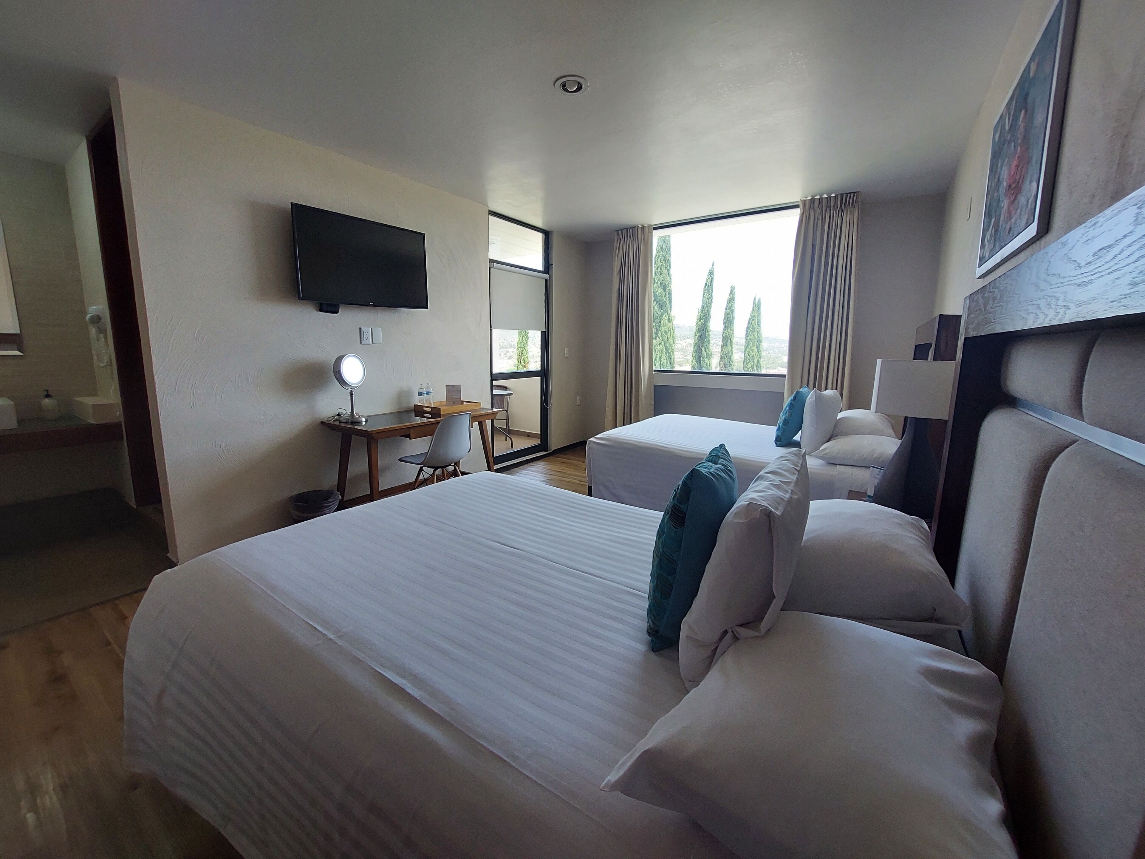 A hotel room with two beds, a desk with a chair, a TV mounted on the wall, and a window with curtains.
