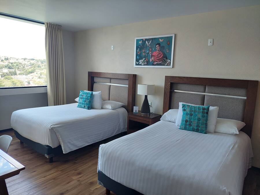Habitación de hotel con dos camas, una mesita de noche con lámpara, un cuadro en la pared y una ventana con cortinas.