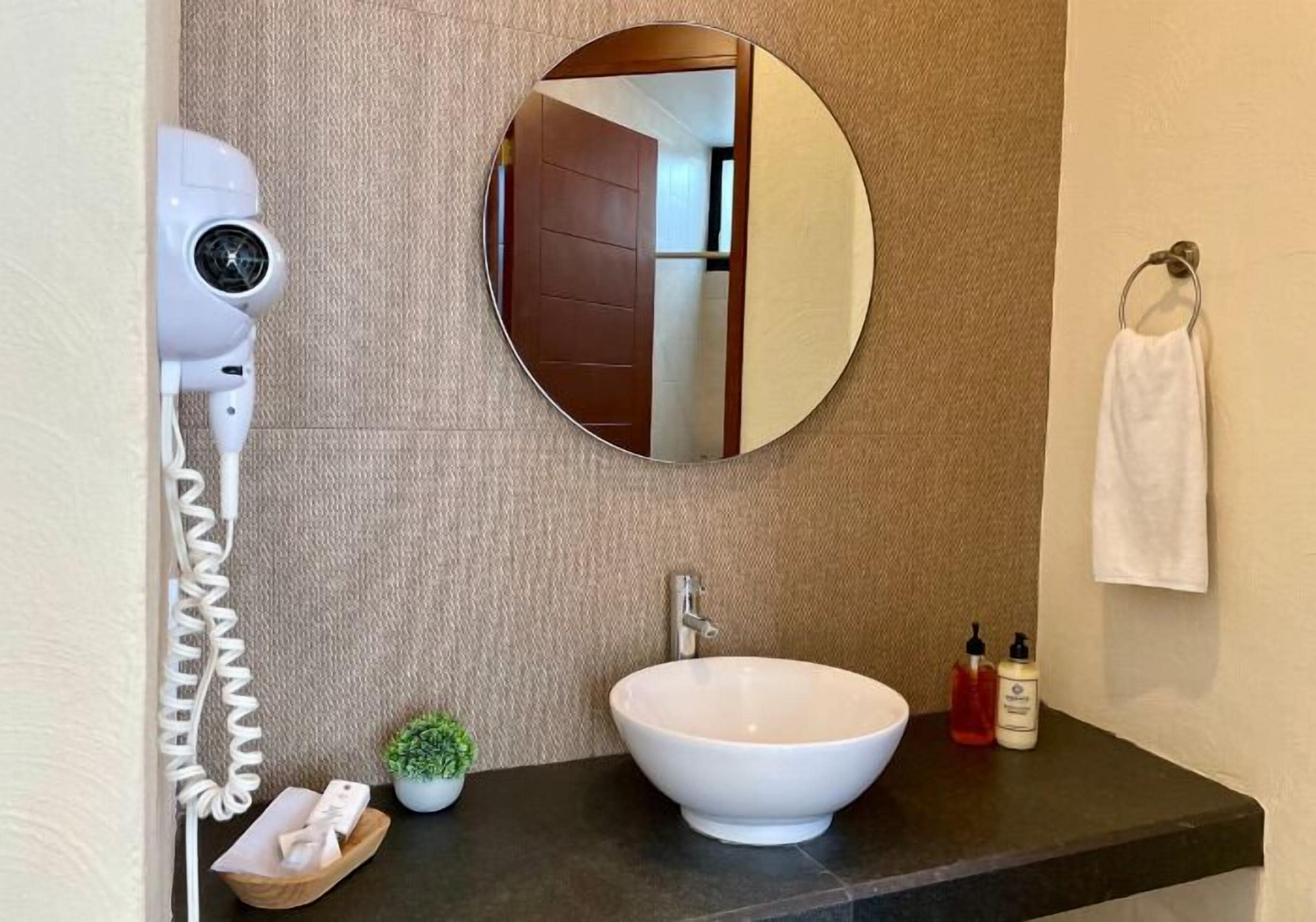 A bathroom with a round mirror, a white sink, a hairdryer, a small potted plant, and two bottles of soap.