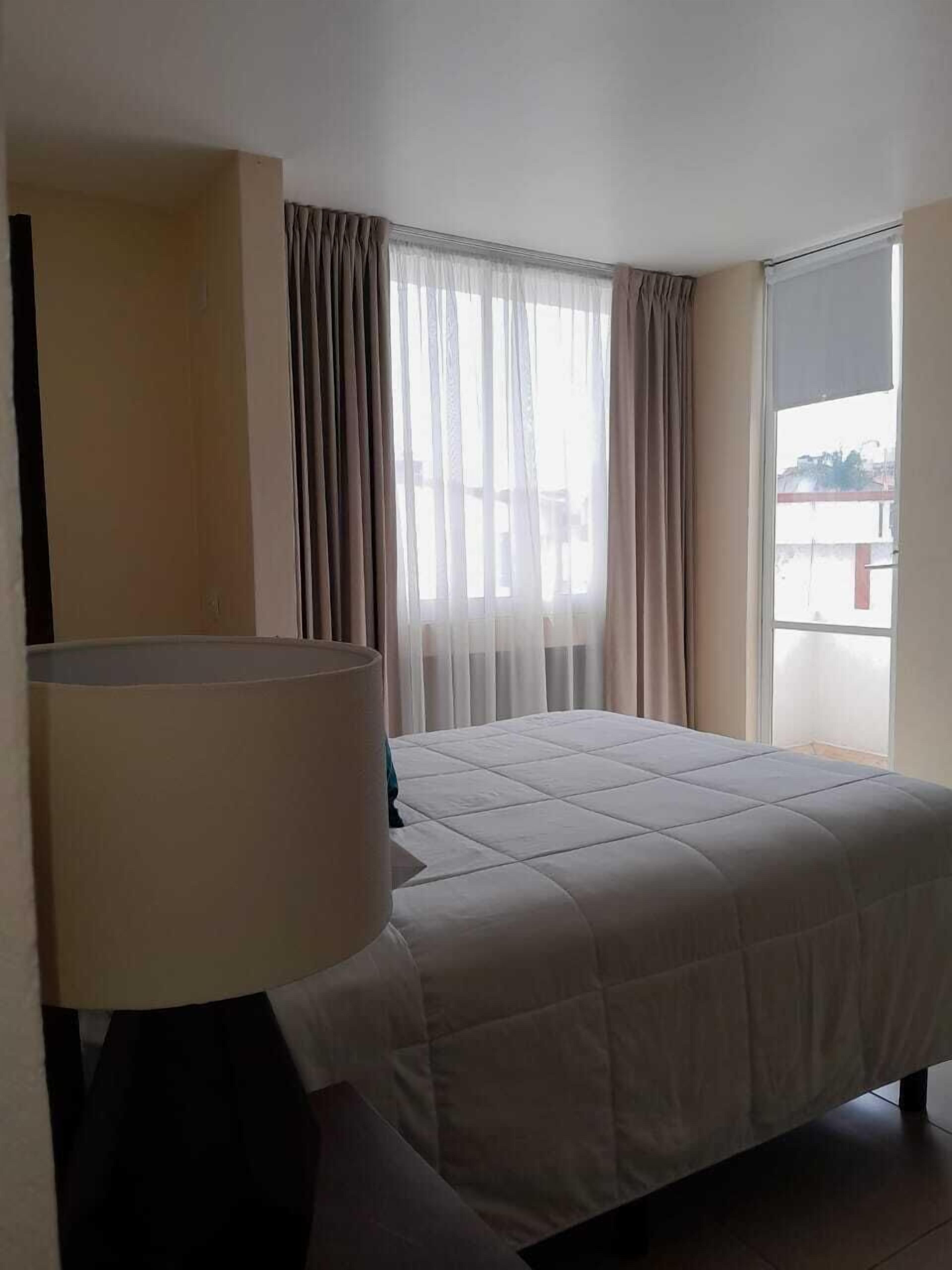 Habitación de hotel con cama, lámpara, ventana con cortinas y vista al exterior.