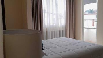 Habitación de hotel con cama, lámpara, ventana con cortinas y vista al exterior.
