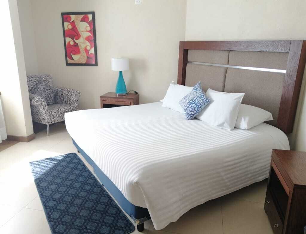 A hotel room with a large bed, a nightstand with a lamp, a patterned armchair, and a wall art piece.