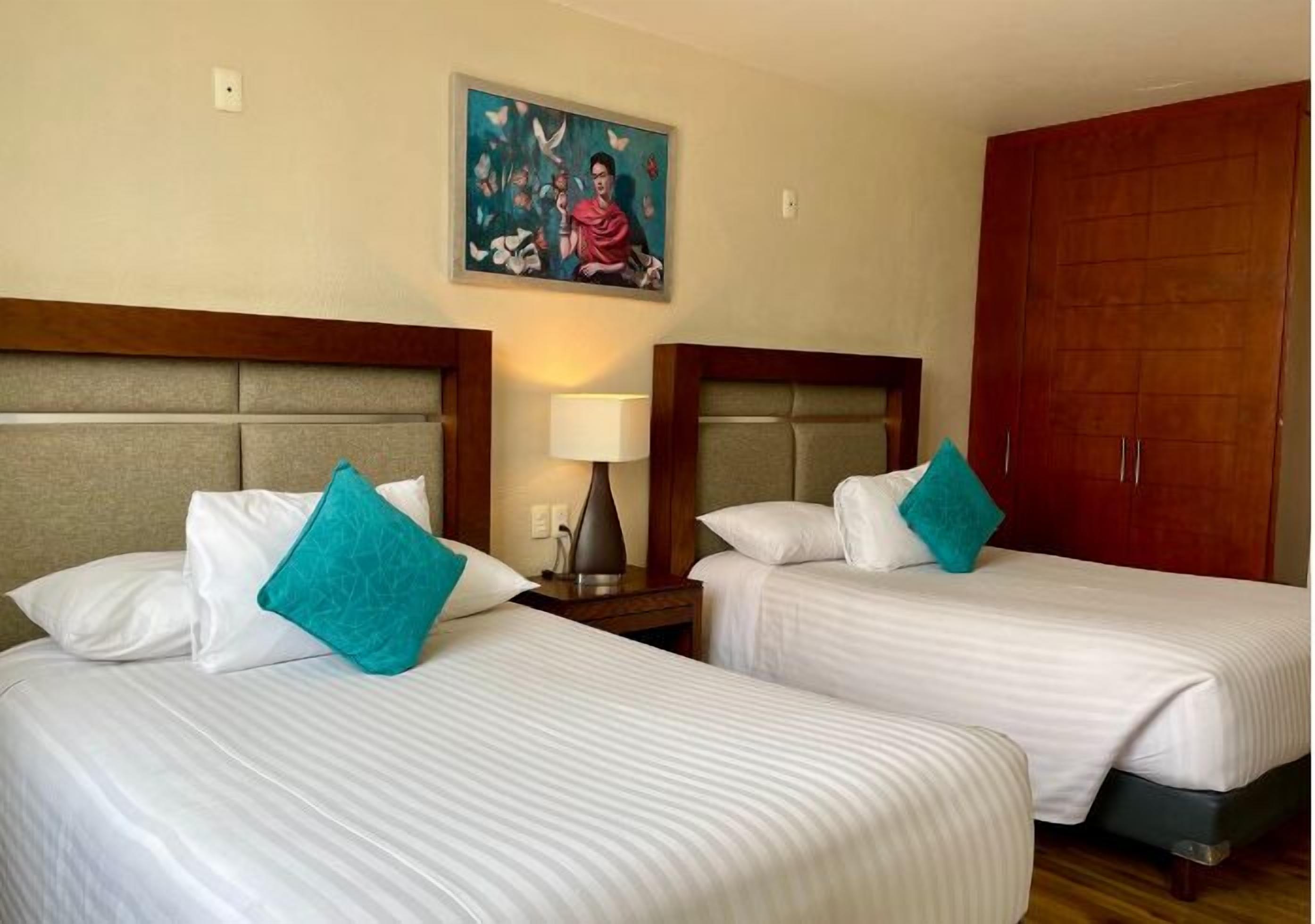 A hotel room with two beds, each with white bedding and teal pillows, a nightstand with a lamp, a painting on the wall, and a wooden wardrobe.