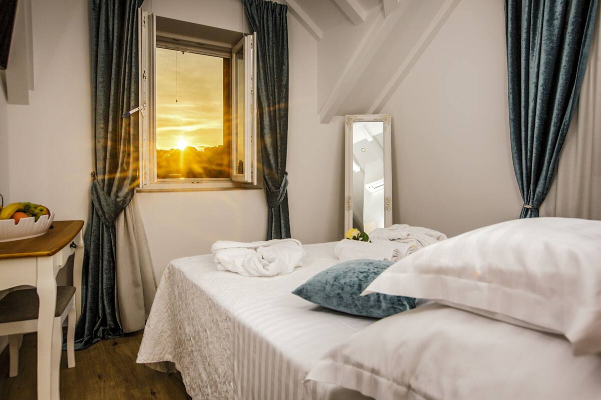 A hotel room with a bed, a window with curtains, a bedside table with a fruit bowl, and a mirror on the wall.