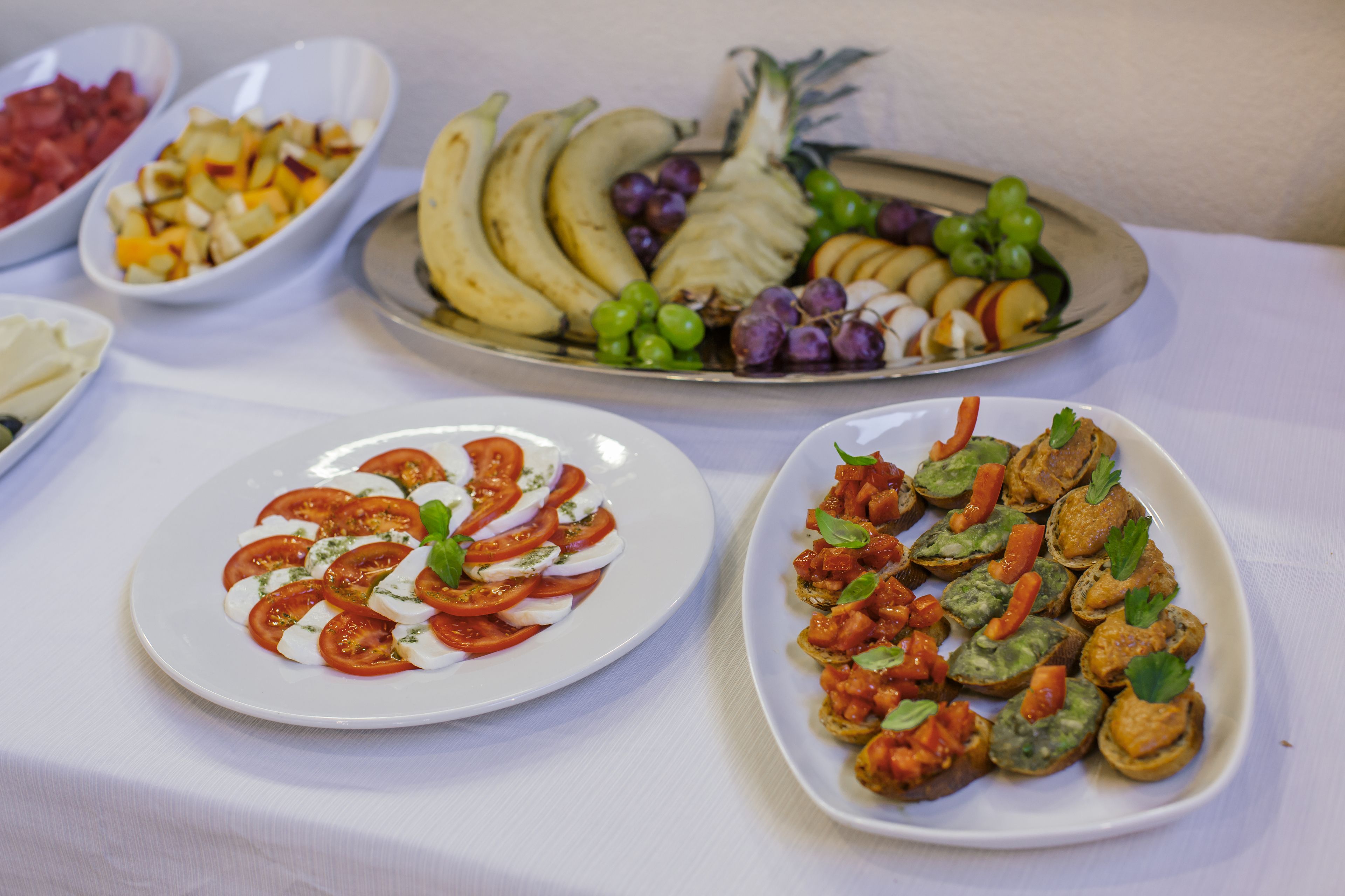 Une table sur laquelle on trouve divers aliments, notamment une assiette de tomates et de mozzarella en tranches, une assiette de bruschettas et une assiette de bananes et de raisins.