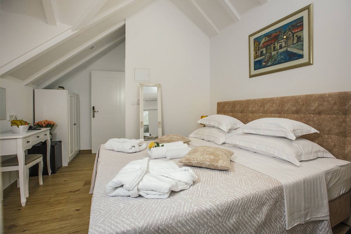 A bedroom with a bed, pillows, a bedside table, a mirror, and a painting on the wall.