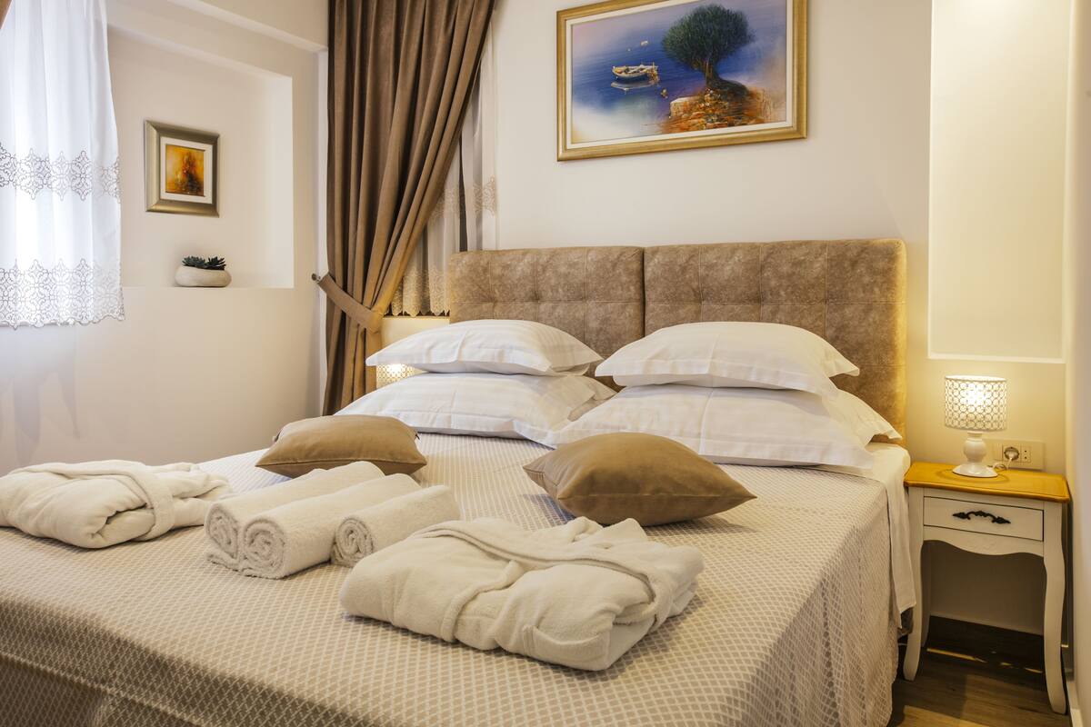 A neatly made bed with white linens and rolled towels, a bedside table with a lamp, and a painting on the wall.