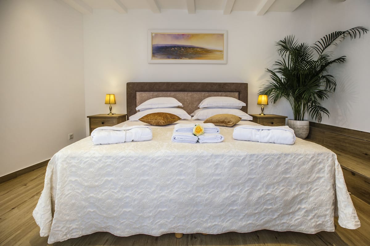 A neatly made bed with a white bedspread, two bedside tables with lamps, a large potted plant, and a painting on the wall.