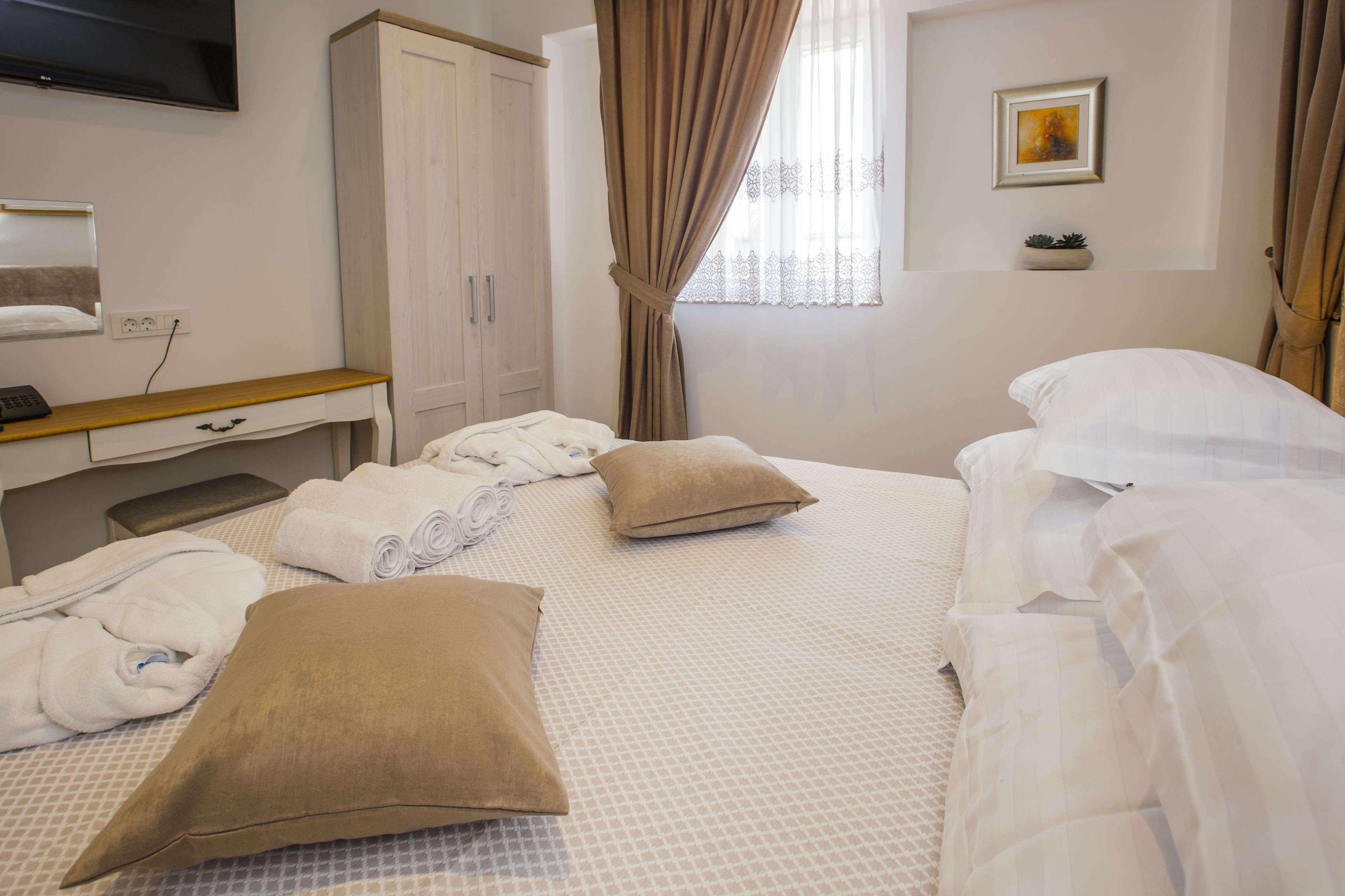 A hotel room with a bed, pillows, a desk with a computer, a TV, and a window with curtains.