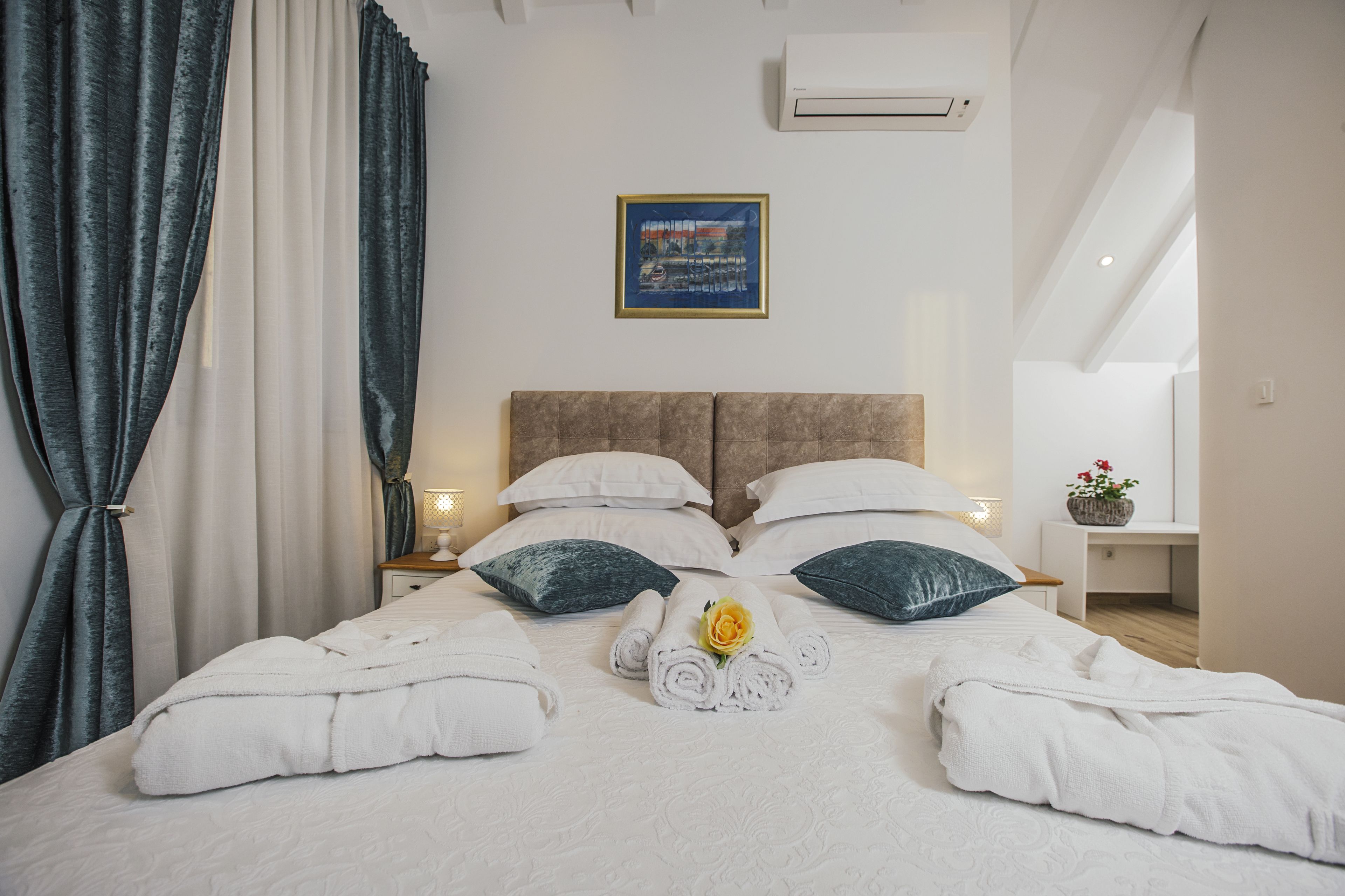 A neatly made bed with white linens, two pillows, and a rolled towel with a flower.