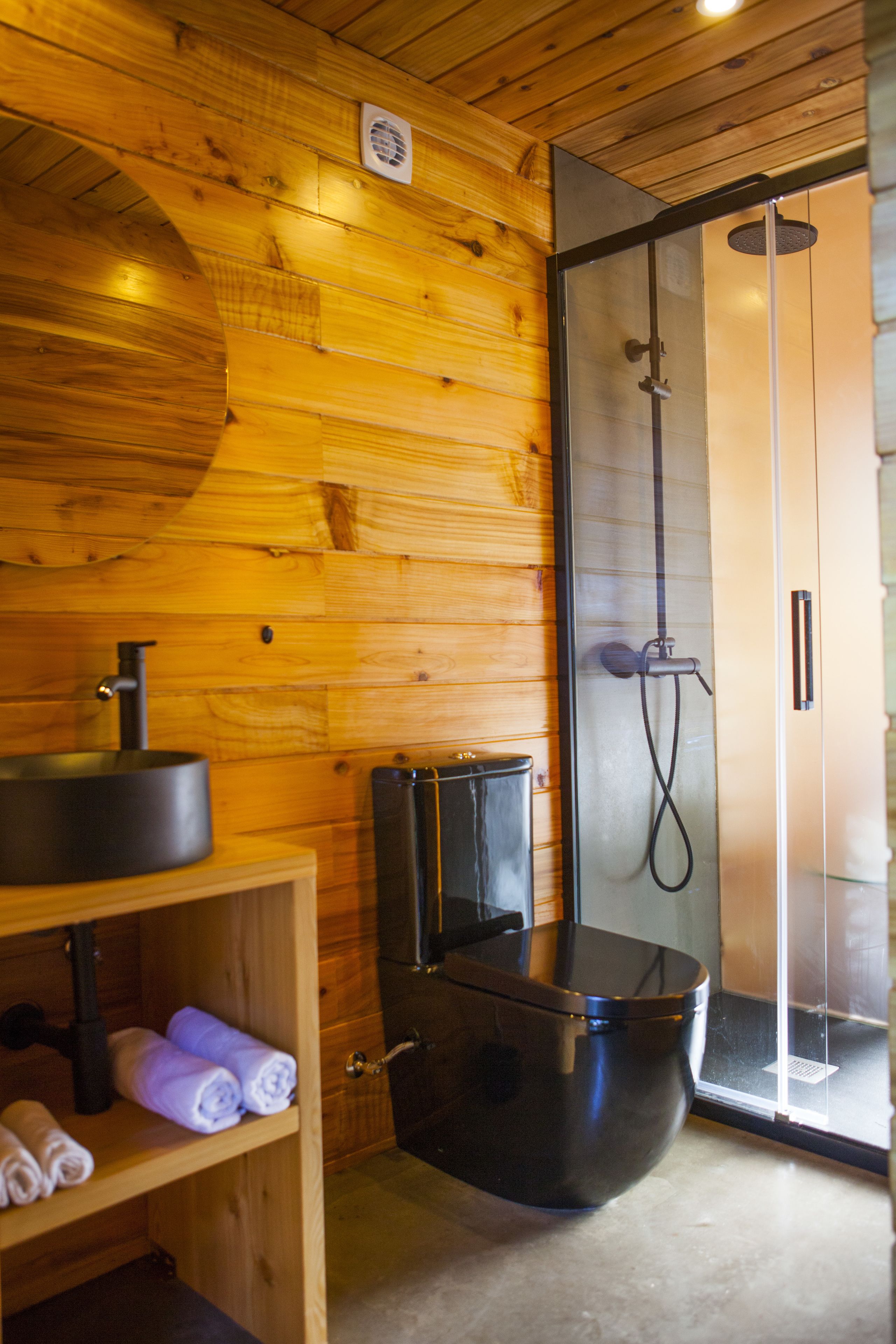 Een houten interieur met een douche in een glazen cabine, een zwart toilet en een plank met handdoeken.
