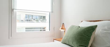 Ein Schlafzimmer mit Bett, Nachttisch, Lampe und einem Fenster mit Blick auf ein Gebäude.