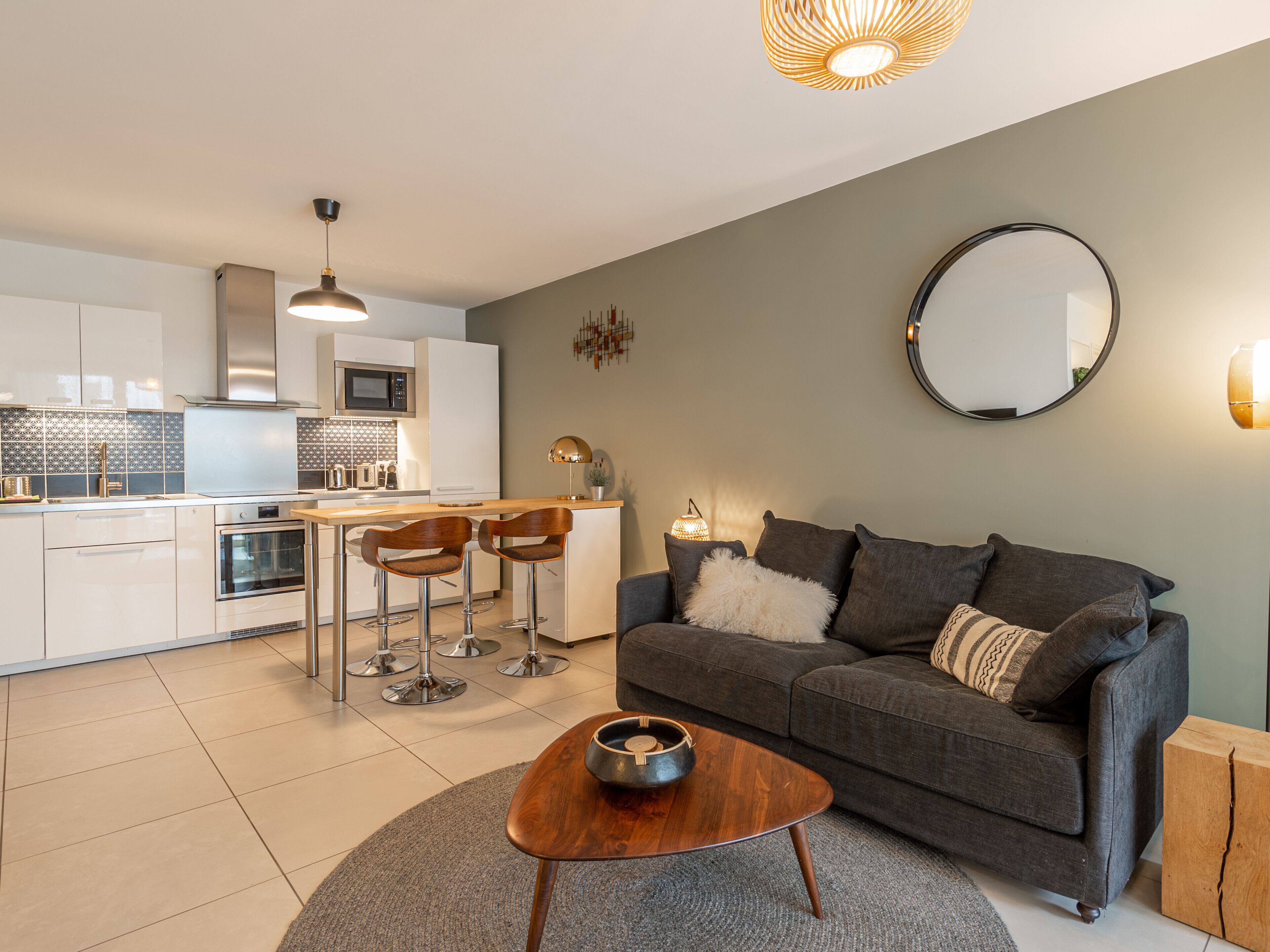 A modern kitchen with white cabinets, a microwave, and a stove. There is a dining area with a wooden table and two bar stools. A gray sofa is against the wall, and a round mirror hangs above it.