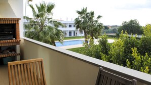 A balcony with a view of a swimming pool, palm trees, and a building in the background. - Condomínio Vilita, T1-Lt-5,F Pêra-Silves (Pêra)