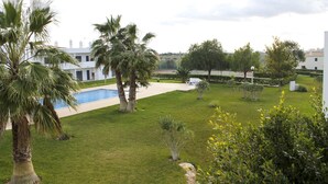 A pool area with palm trees, a tennis court, and a white building in the background. - Condomínio Vilita, T1-Lt-5,F Pêra-Silves (Pêra)