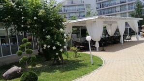 A covered outdoor seating area with tables and chairs, surrounded by greenery and a building in the background. - Charis Cozy Family Apart. at Sun Village  - ONLY 10 mins Walk to the Beach (Bourgas)