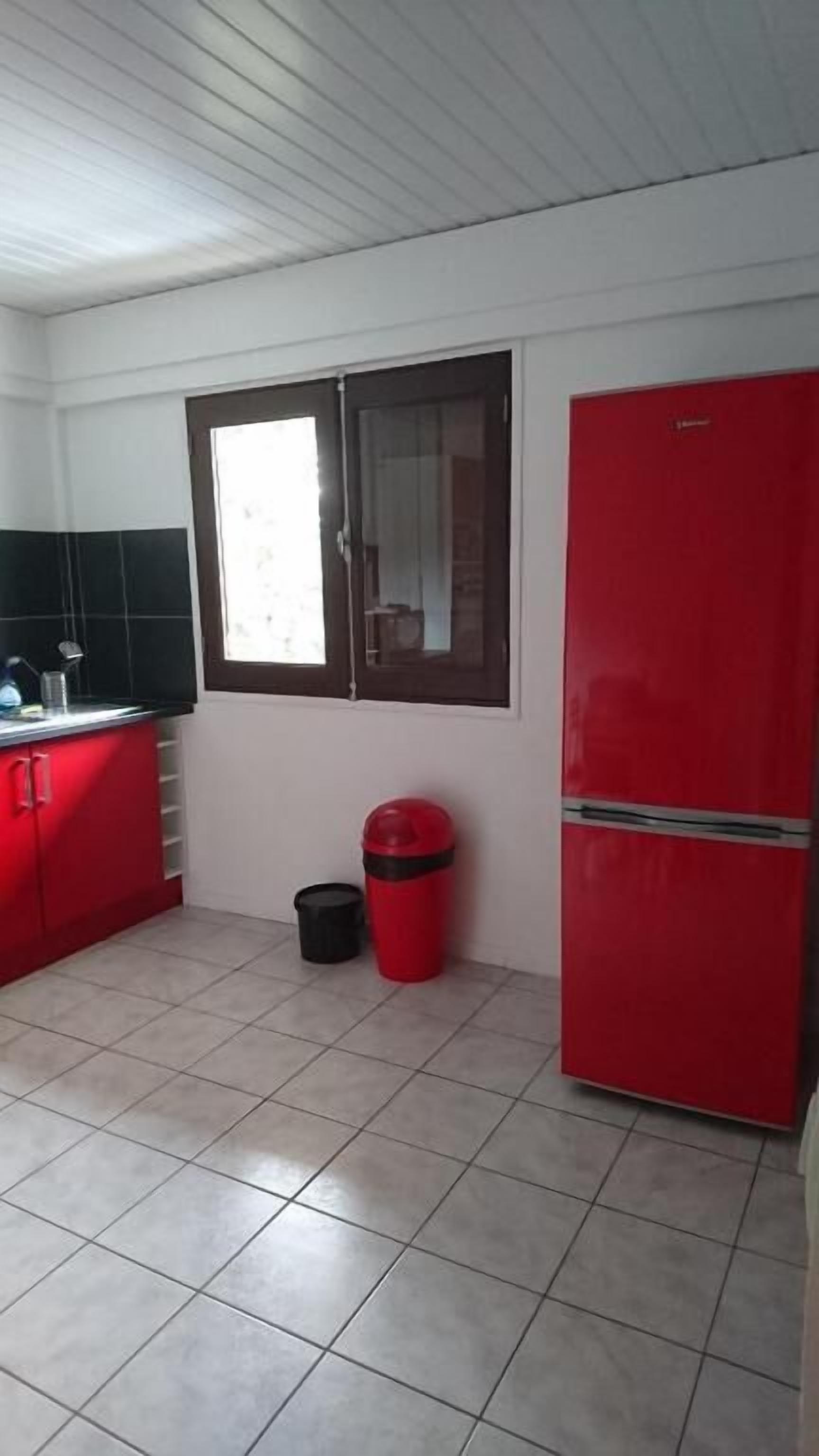 A kitchen with a red refrigerator, a window with a brown frame, and a trash bin.