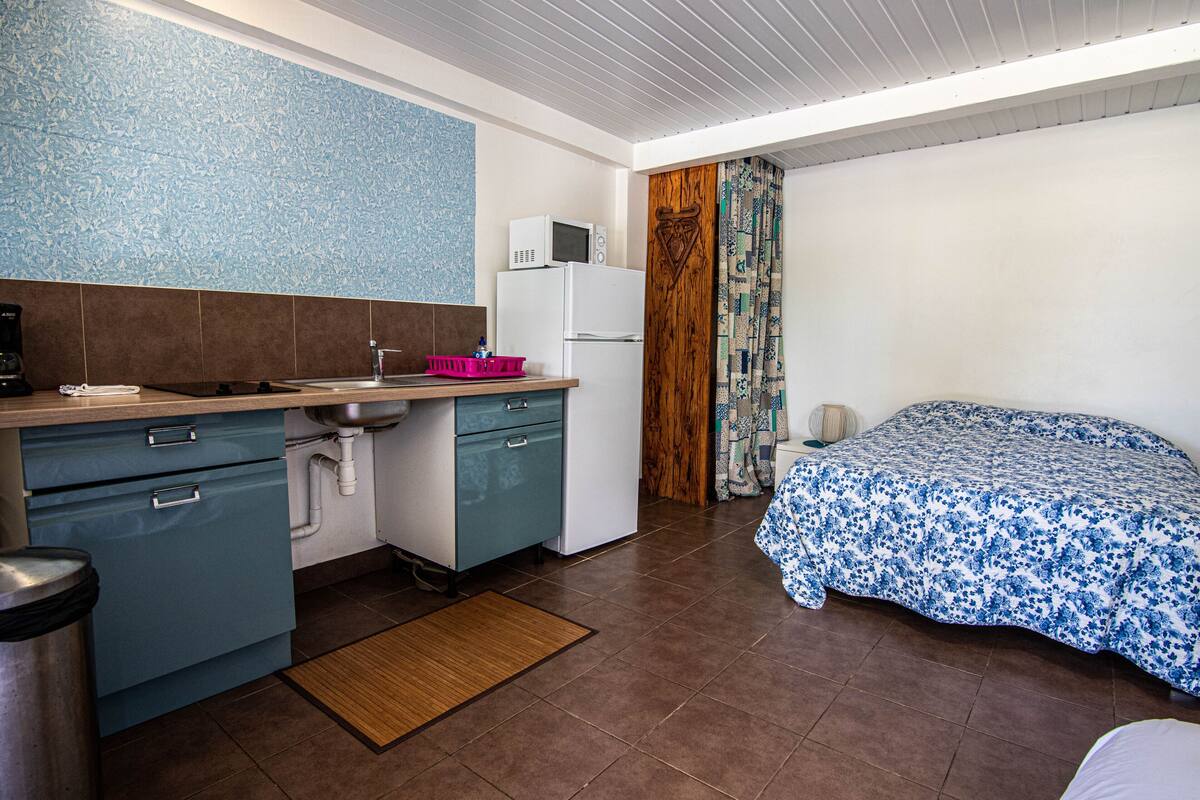 Une cuisine compacte équipée d’un micro-ondes, d’un évier et de placards, un petit coin repas avec une table et des chaises, et une chambre avec un lit et des rideaux.