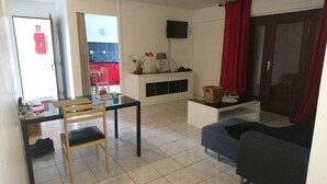 Un salon comprenant une table à manger, un canapé, un téléviseur fixé au mur et une cuisine visible à travers une porte ouverte.