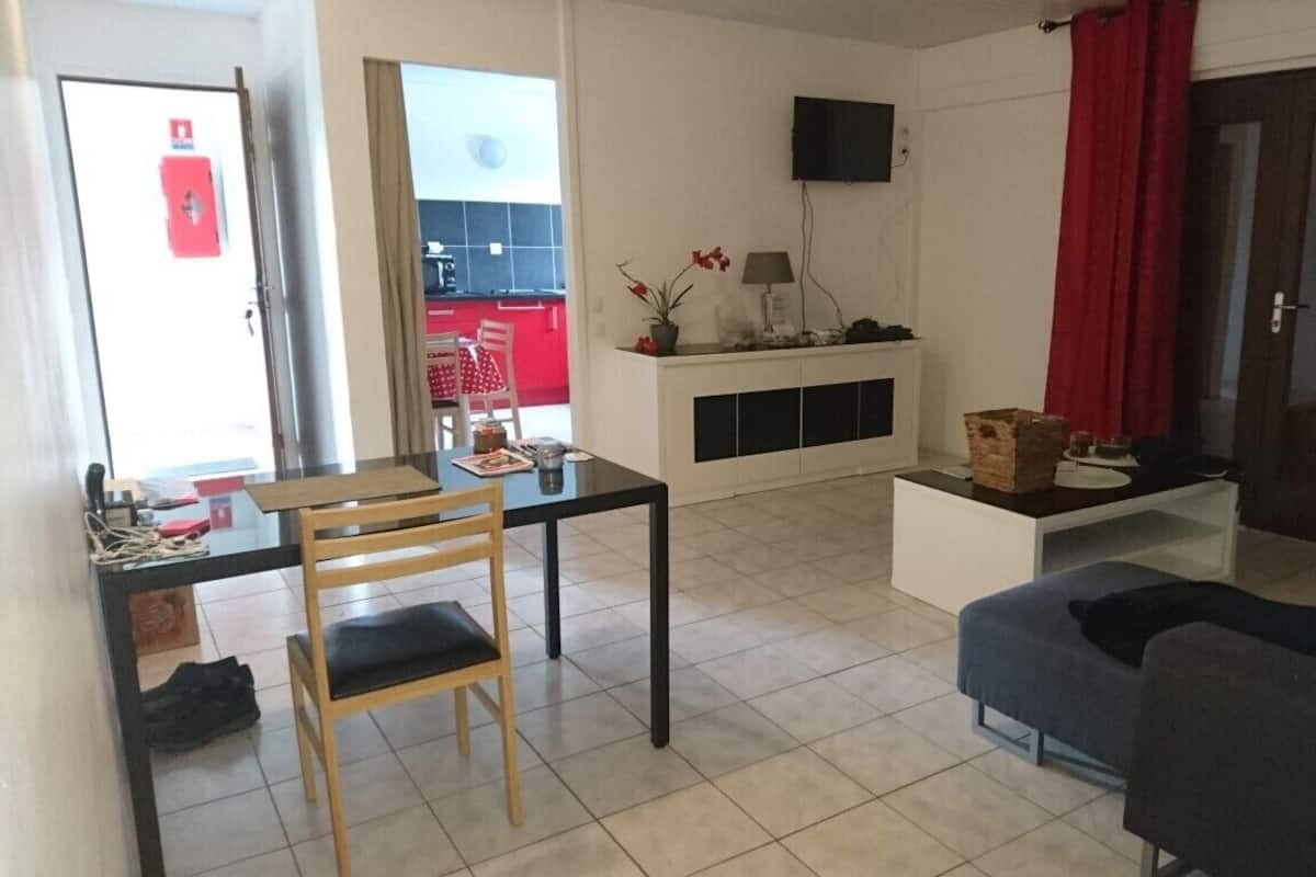 Un salon comprenant une table à manger, un canapé, un téléviseur fixé au mur et une cuisine visible à travers une porte ouverte.