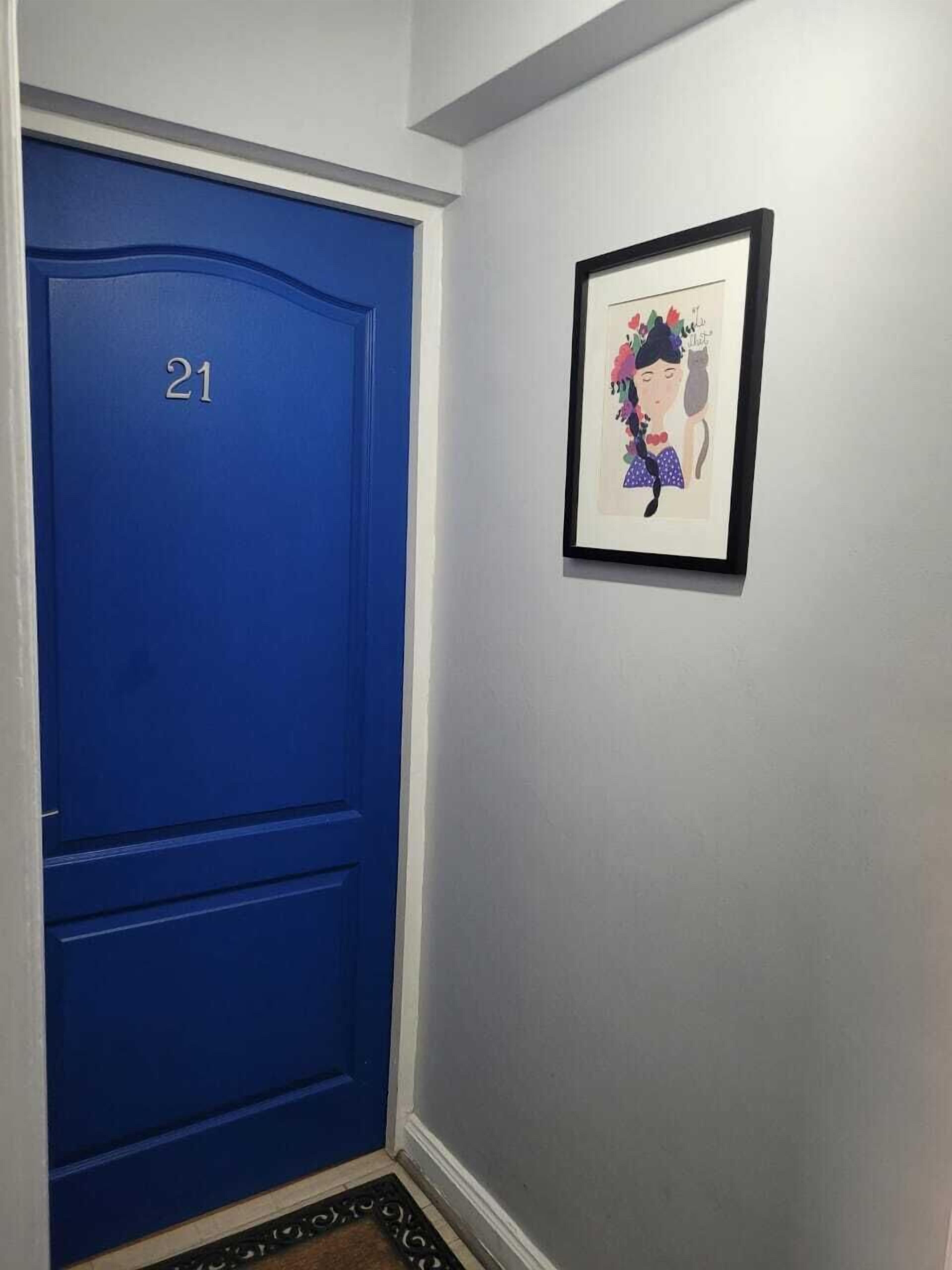 Una puerta azul con el número 21, un cuadro en la pared y un felpudo en la entrada.