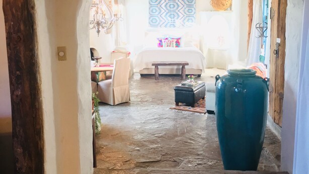 A rustic hallway leading to a bedroom with a bed, a chandelier, and a patterned wall hanging.