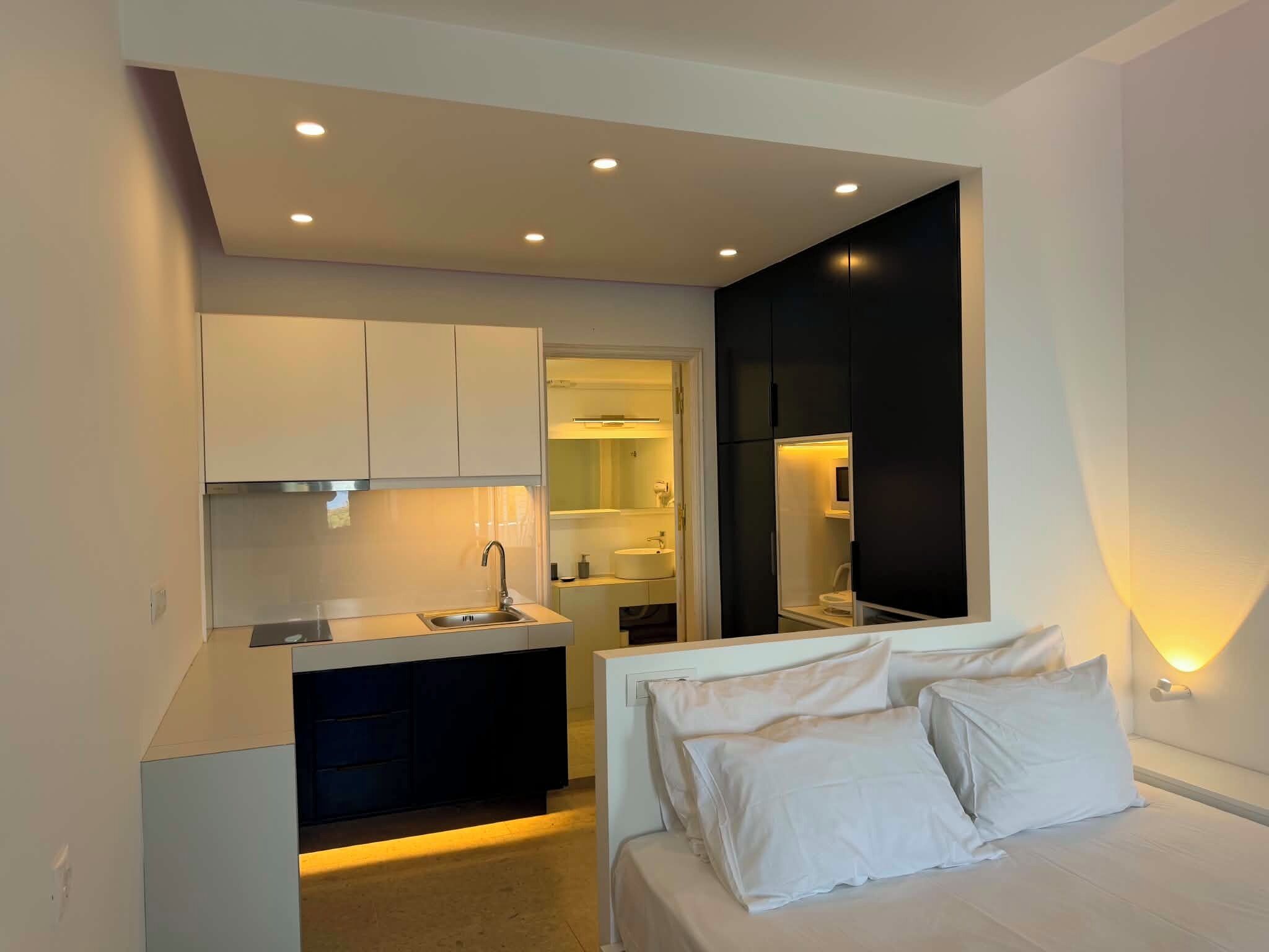 A compact bedroom with a bed, a small kitchenette, and a bathroom visible through an open door.