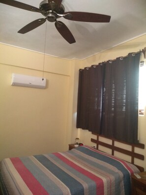 Quarto com cama, ventilador de teto, ar condicionado e janela com cortinas.